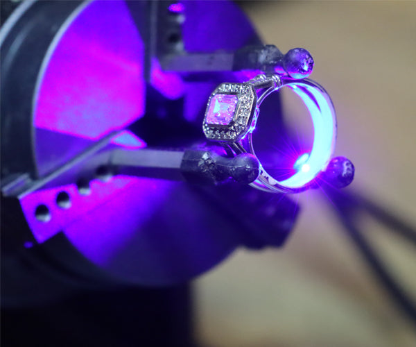 Gros plan sur une bague sertie de pierres précieuses fixée dans une machine de gravure laser, illuminée par un faisceau violet lors de la personnalisation en atelier chez Flamme en rose.