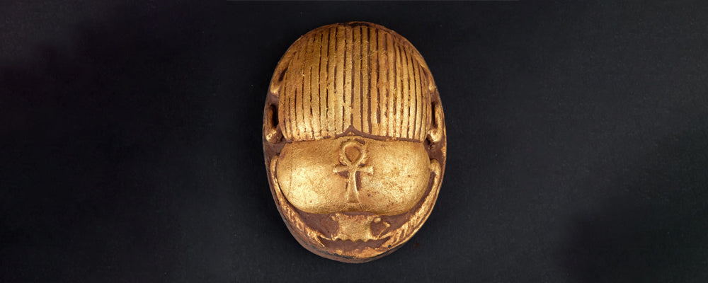 Amulette égyptienne dorée en forme de scarabée, ornée du symbole Ankh gravé sur le dos, symbole de vie éternelle dans l’Égypte ancienne.