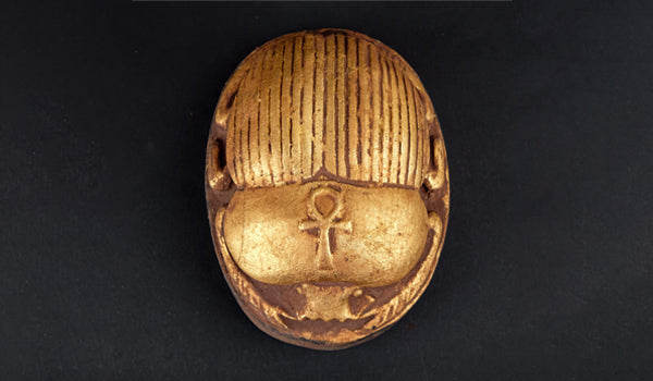Amulette égyptienne dorée en forme de scarabée, ornée du symbole Ankh gravé sur le dos, symbole de vie éternelle dans l’Égypte ancienne.