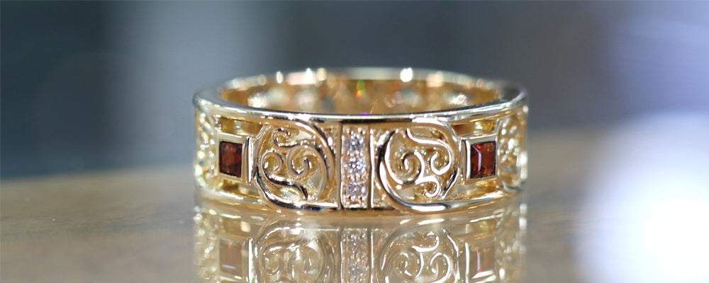 Bague en or jaune photographiée sur une table en verre, ornée de grenats carrés et de petits diamants. Le corps ajouré présente un motif décoratif symétrique de style celtique, intégré dans une structure rectangulaire régulière.