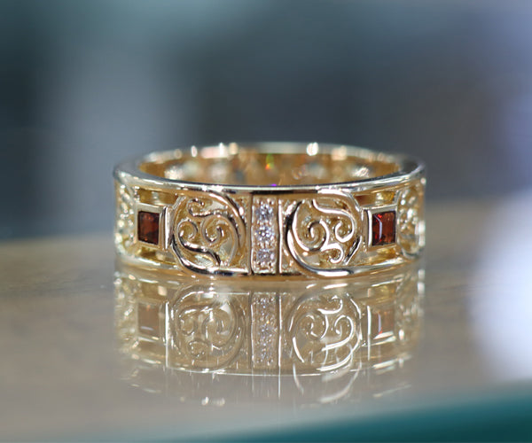 Bague en or jaune photographiée sur une table en verre, ornée de grenats carrés et de petits diamants. Le corps ajouré présente un motif décoratif symétrique de style celtique, intégré dans une structure rectangulaire régulière.