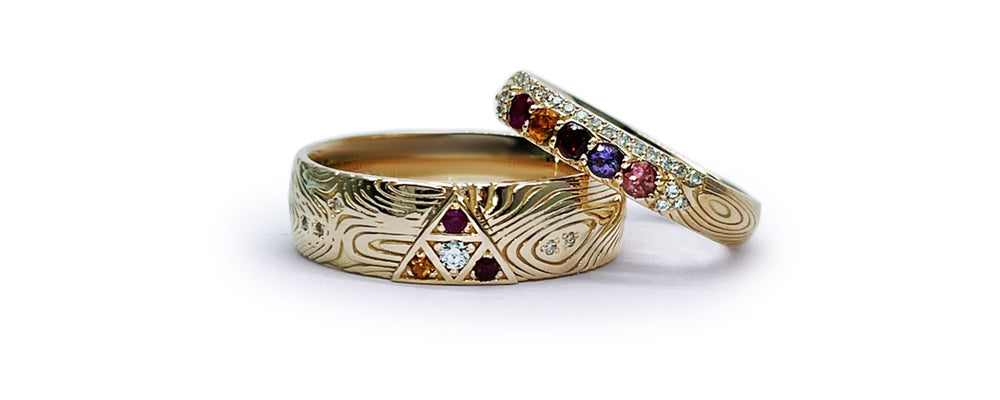 Ensemble de bagues en or jaune faites sur mesure. L’anneau homme, plus large, présente un motif triangulaire serti de diamant, grenat et rubis, sur un corps de bague texturé de style damas. L’anneau femme est pavé de pierres naturelles : grenat, spinelle, saphir violet, tourmaline rose et diamants.