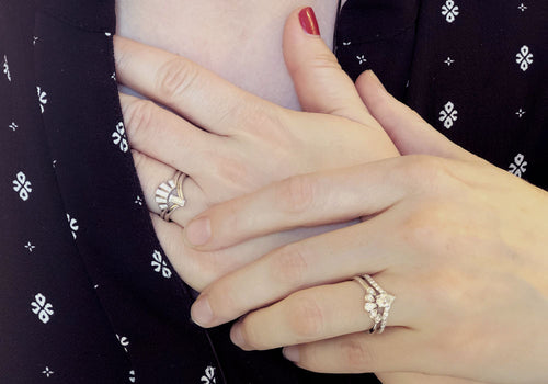 Two white women's hands holding rings on the white gold ring finger.